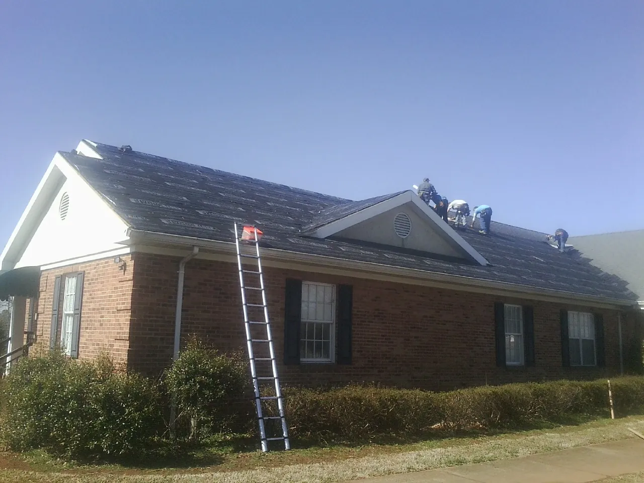 Crew tearing shingles off a brick ranch roof with a ladder set — Metal Roof Repair work by our crew in St Columbans